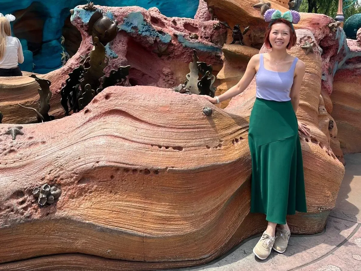 Kristy wearing themed ears at a Disney park, beside a Little Mermaid lagoon set